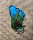 A footprint wooden magnet with a green and blue background featuring an engraved image of bigfoot and a forest.