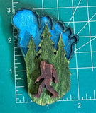 A footprint wooden magnet with a green and blue background featuring an engraved image of bigfoot and a forest on a measuring mat.