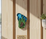 Handcrafted wooden magnet with a nature design and a sasquatch on a wooden surface, accompanied by a tag.