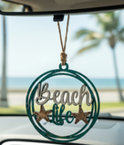 Decorative car hanging with 'Beach Life' text and star designs, hanging from a car's rearview mirror with a beach view in the background.