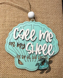 A wooden car charm with a shell design and the text 'Call Me On My Shell' written on it, against a burlap background.