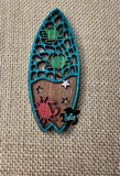 A wooden magnet shaped like a surfboard with a crab and turtle design.