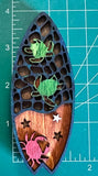 A wooden magnet shaped like a surfboard with a crab and turtle design with measuring mat in background measuring 4 inches