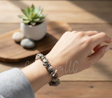 Hand wearing a bracelet on a wooden surface with a plant in the background