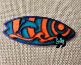 A colorful wooden magnet shaped like a dolphin surfboard with a vibrant blue-and-orange design, placed on a beige textile surface.