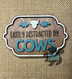A rectangular wooden magnet with an engraved text 'EASILY DISTRACTED BY COWS' surrounded by a blue leafy border, on a burlap background.