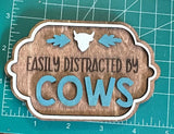 A rectangular wooden magnet with an engraved text 'EASILY DISTRACTED BY COWS' surrounded by a blue leafy border, on a measuring mat background.