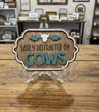 Wooden magnet with 'Easily Distracted by Cows' text on a stand in a store setting.
