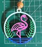 A wooden car charm with a flamingo design, pink, green, and blue colors, with a string for hanging, with a measuring mat background