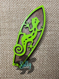 A magnet shaped like a green gecko mounted on a brown surfboard-shaped magnet.