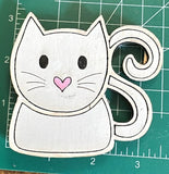 A magnet shaped like a stylized cat with a gray color and a heart on its chest, made from wood on a measuring mat. 