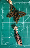 May include: A wooden butterfly charm with a brown and black woodgrain finish, a white flower charm, and a brown feather charm with a dark brown woodgrain finish. The charms are strung together with a tan twine.
