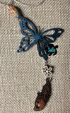 May include: A teal blue butterfly charm with a white flower and a brown wooden feather charm hanging from it. The charms are strung on a tan twine. The charms are on a burlap background.