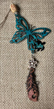 May include: A turquoise butterfly charm with a white flower and a brown wooden feather charm hanging from it. The charms are strung on a tan twine. The charms are on a burlap background.