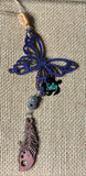 May include: A blue butterfly charm with a brown feather charm and a silver flower charm hanging from a twine string with a wooden bead. The charms are hanging on a burlap background. 