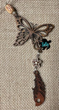 May include: Wooden butterfly charm with a brown wood flower and a brown wood feather charm. The charms are attached to a brown wood bead with a rope. The charms are on a burlap background. 