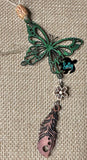 May include: A green butterfly charm with a brown wooden bead and a flower charm hanging from it. The butterfly has a detailed design and is made of wood. The charm is attached to a tan cord.