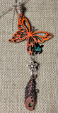 May include: Orange wooden butterfly with a white flower and a brown wooden feather charm hanging from it. The charm is attached to a tan cord. The butterfly and feather are both intricately carved. The background is a brown burlap fabric.