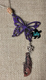 May include: A purple butterfly charm with a wooden flower and feather charm hanging from it. The charms are attached to a brown twine string with a wooden bead.