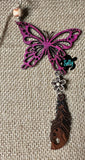 May include: A pink wooden butterfly with a brown wooden feather charm and a white flower charm. The charms are attached to a silver chain. The butterfly and charms are hanging from a brown twine string. The background is a brown burlap fabric.