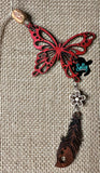 May include: A red wooden butterfly with a white flower and a brown wooden feather charm. The butterfly has a delicate, intricate design and is hanging from a brown twine string. The charm is attached to the butterfly with a silver chain. The background is a brown burlap fabric.
