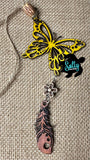 May include: A yellow butterfly charm with a white flower and a brown wooden feather charm hanging from it. The charms are strung on a tan twine. The charms are on a burlap background.