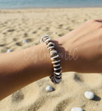 A handmade bracelet featuring alternating natural brown coconut husk discs and white puka shell beads, worn on a wrist at a sandy beach