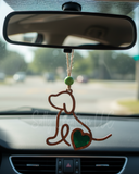 Handmade wooden dog silhouette car charm with a green heart inlay, hanging from a rearview mirror by tan twine inside a car.
