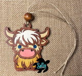 May include: A wooden ornament of a Highland Cow with brown, white, and yellow fur. The cow has blue eyes and a pink nose. The ornament is hanging from a brown twine string with a brown wooden bead at the top. 
