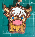 May include: A wooden ornament shaped like a Highland cow with brown and white fur, a pink nose, and blue eyes. The ornament has a hole at the top for hanging.