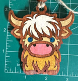 May include: A wooden ornament shaped like a Highland cow with brown and white fur, a pink nose, and blue eyes. The ornament has a hole at the top for hanging.