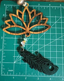 A wooden car charm with a lotus flower design, featuring hanging  wooden feather and strings and a small beaded ornament laying on a measuring mat