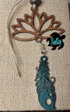 A wooden car charm with a lotus flower design, featuring hanging teal wooden feather and strings and a small beaded ornament.