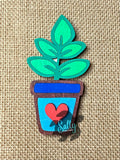 A hand-made magnet shaped like a green plant in a brown pot with a heart symbol, placed on a burlap background.
