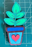 A hand-made magnet shaped like a green plant in a brown pot with a heart symbol, placed on a measuring mat.