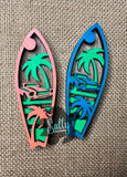 Two wooden magnets with a tropical classic car surfboard design, one in orange and one in blue, placed on a burlap fabric surface.