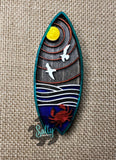 A hand-made wooden magnet with a surfboard shape, featuring a sunset, moon, seagulls, and a crab engraving.