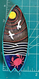 A hand-made wooden magnet with a surfboard shape, featuring a sunset, moon, seagulls, and a crab engraving on a meaasuring mat. 