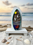 Decorative surfboard with ocean-themed design on a sandy beach with shells and driftwood.