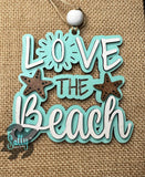 May include: A wooden sign with the words "LOVE THE BEACH" in white and teal, featuring two starfish. The sign has a string for hanging and a white bead at the top. 