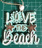 May include: A wooden beach-themed ornament with the words "LOVE THE BEACH" in white, brown, and light blue. The ornament features two starfish and a sun design. It is suspended by a twine string and a white wooden bead.