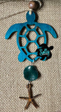 A wooden car charm in the shape of a sea turtle with a turquoise acrylic bead, hanging from a string.