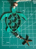 A wooden car charm in the shape of a sea turtle with a turquoise patterned shell, hanging from a string laying on a measuring mat.