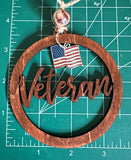 A wooden car charm with the word 'Veteran' engraved on it, alongside an American flag, hanging from a string laying on a measuring mat