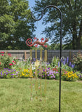 Hand-painted red and green gecko wind chime with golden glass prisms and a sun charm hanging from a black metal shepherd's hook in a lush flower garden.