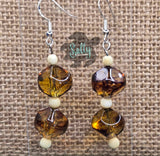 A pair of square-shaped earrings with a grainy, golden brown pattern and white accents displayed against a burlap background.