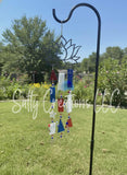 Handcrafted black metal lotus flower wind chime with red, white, and blue stained glass pieces hanging on a shepherd's hook in a sunny garden