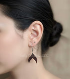Close-up of a woman wearing a dark, handcrafted wooden mermaid tail earring from Salty Creations LLC. The earring features a natural wood grain and hangs from a silver hook.